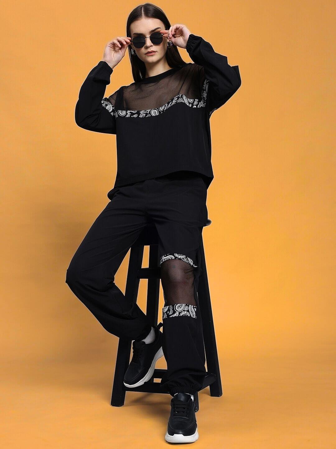 Model wearing WOMEN'S YESMIN RELAXED FIT TOP BLACK with sunglasses on a stool against a yellow background