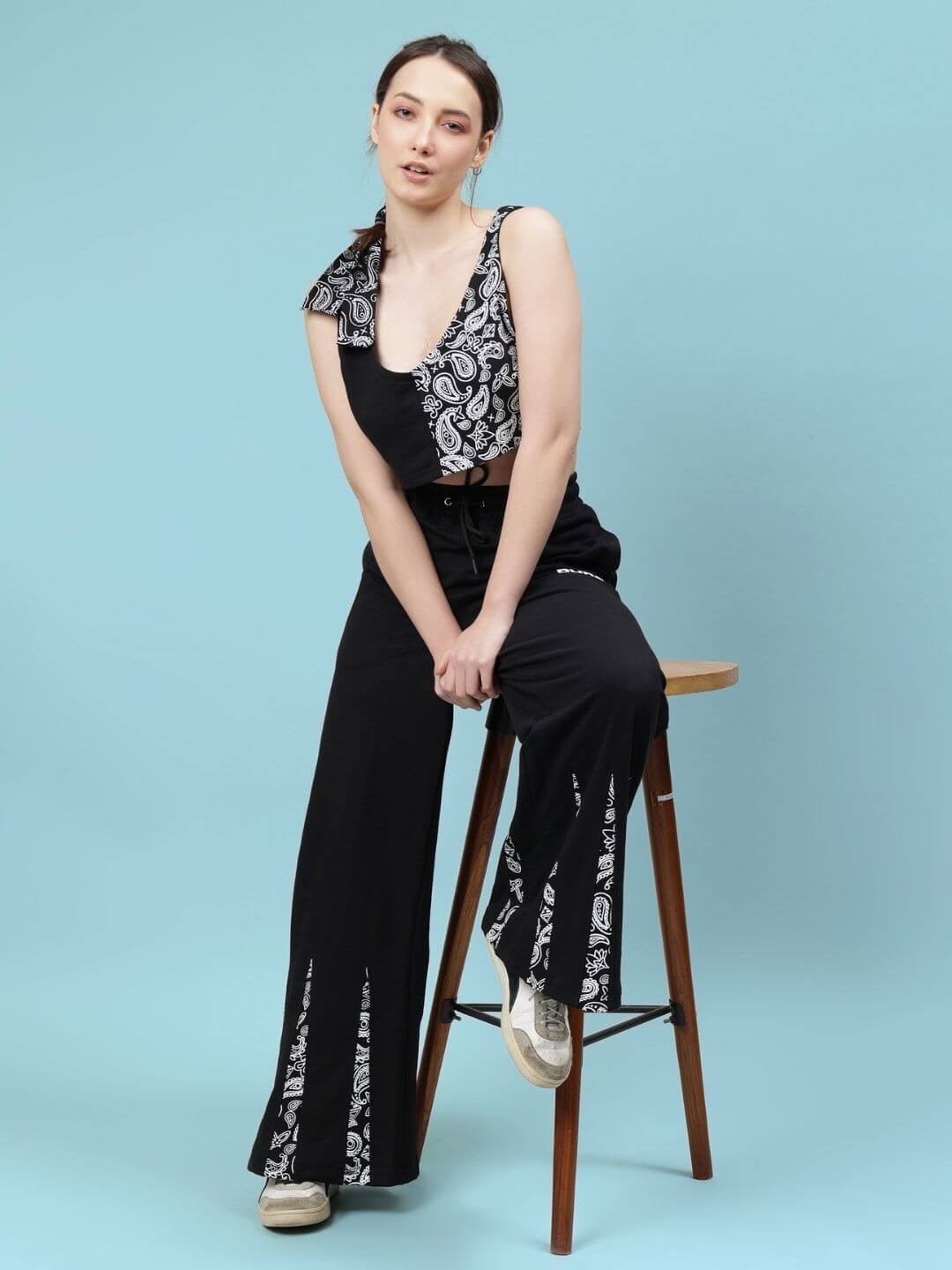 Woman wearing black Ziya co-ord set with paisley design, sitting on stool against blue background, showcasing fashion outfit.