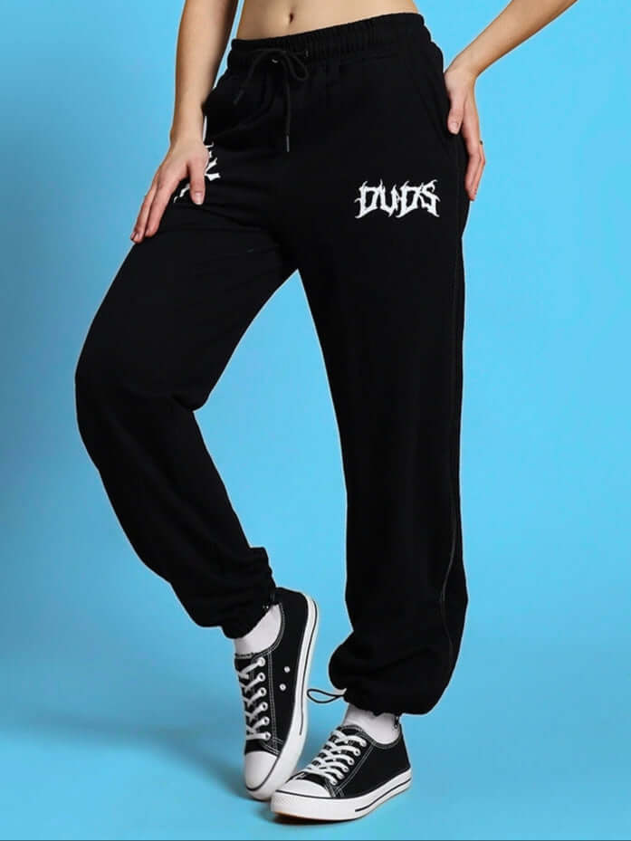 Woman wearing black DUDS joggers with zipper panels, leaning on a blue wall. Casual style for women, customizable and made to order.
