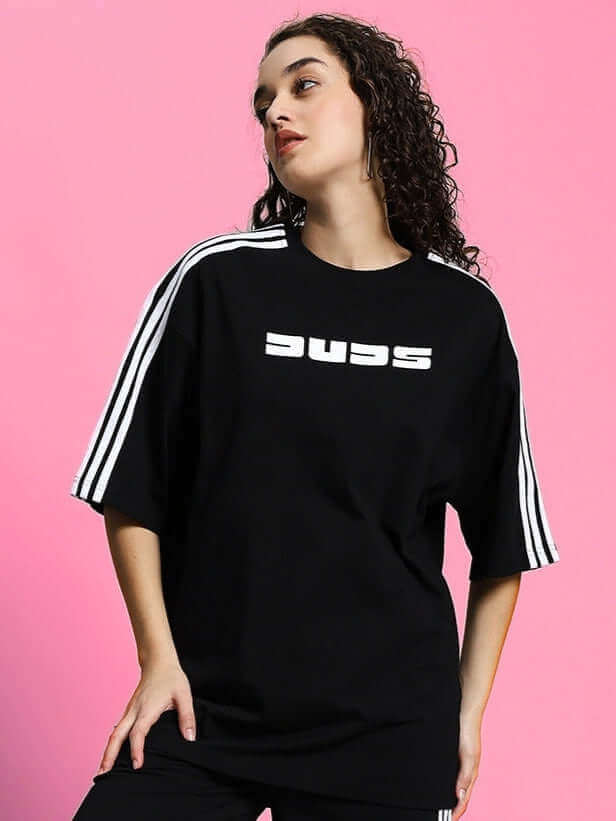 Woman wearing black oversized parka T-shirt with white stripes on sleeves, standing against pink background.
