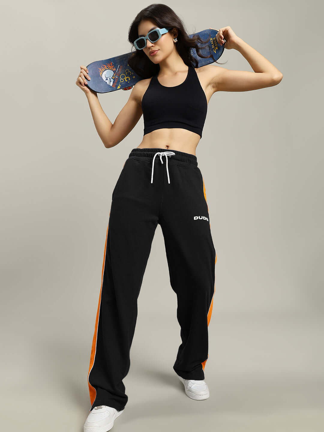 Woman posing in black Sanford Stripe Joggers with orange accents, holding a skateboard, wearing a black crop top and sunglasses.