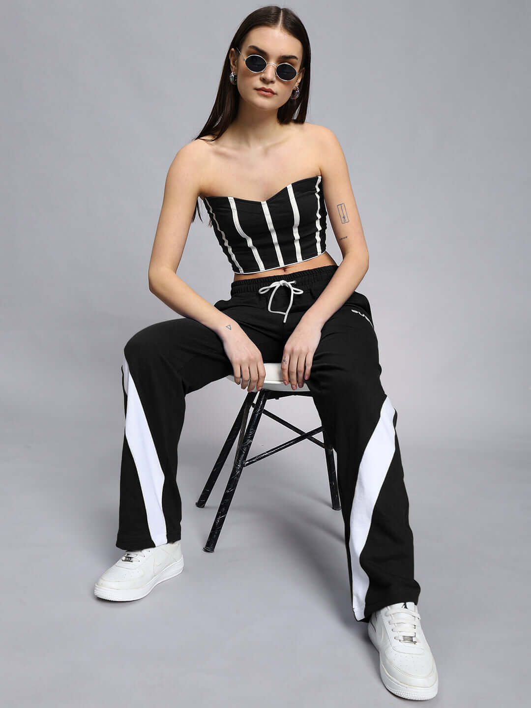 Woman wearing black and white Celestine relaxed fit joggers, seated on a chair, styled with a striped crop top and sunglasses.