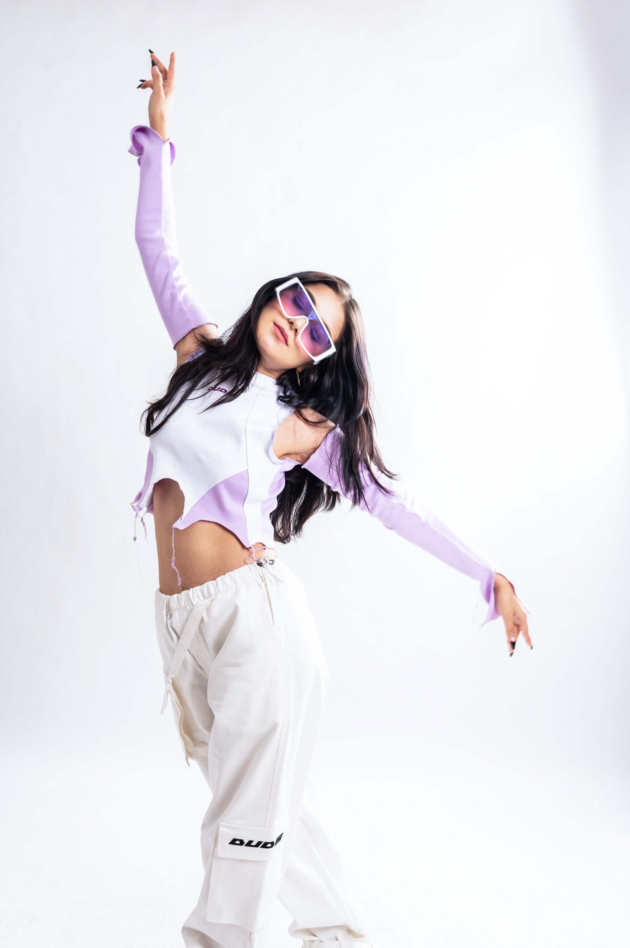 Model wearing a women's color block crop top with long sleeves, paired with white pants, posing energetically. Fashionable and trendy style.