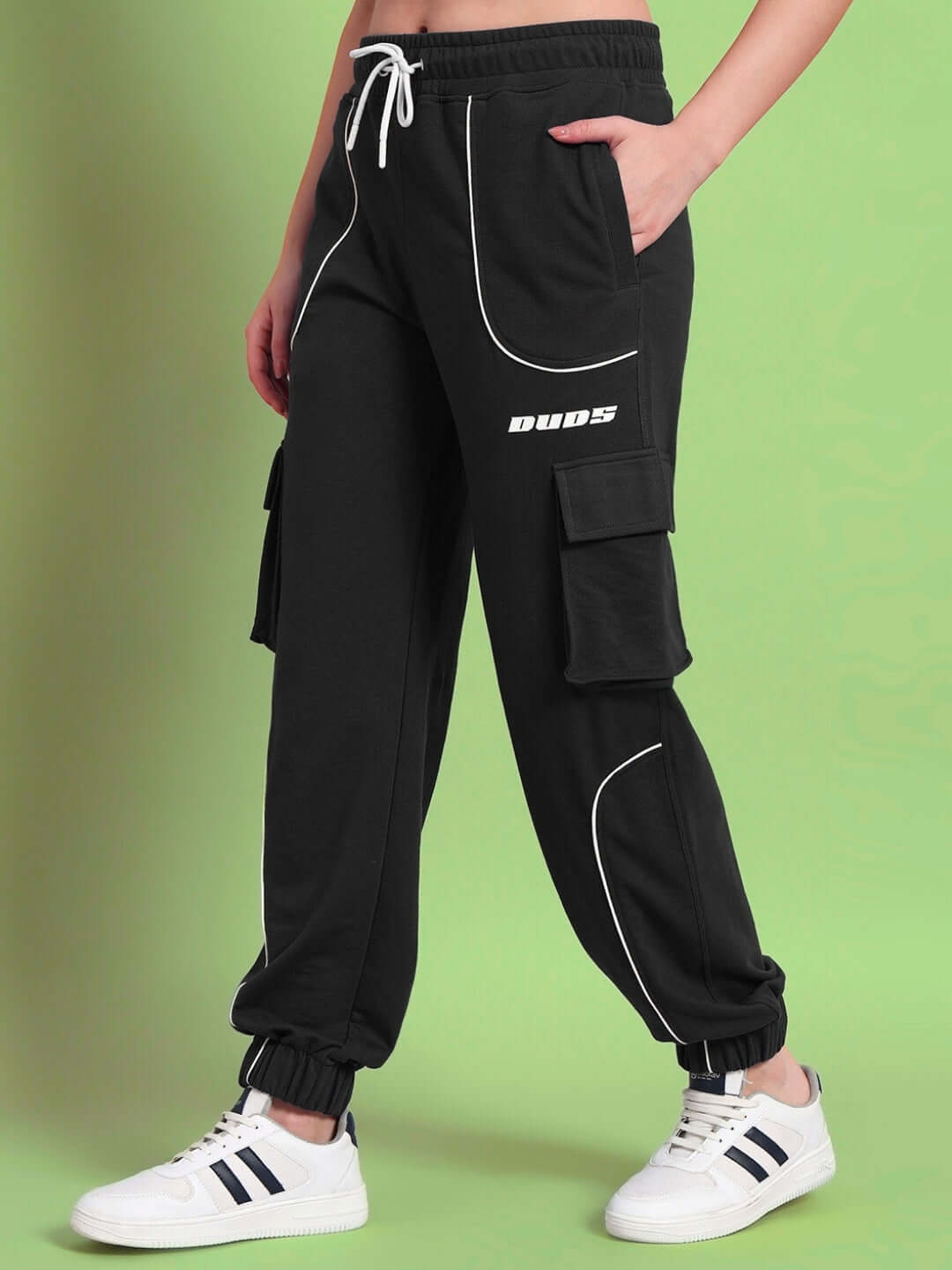 Women's black relaxed-fit joggers by DUDS, featuring cargo pockets and white piping, styled with white sneakers on a green background.