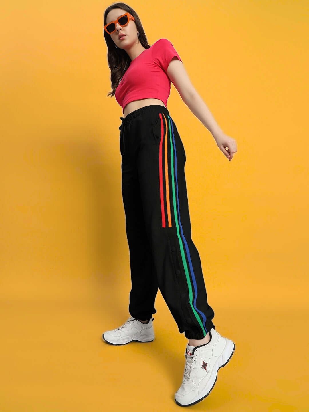 Model wearing Women's DUDS Prism Colorful Stripe Joggers in black with vibrant stripes, paired with white sneakers against a yellow background.