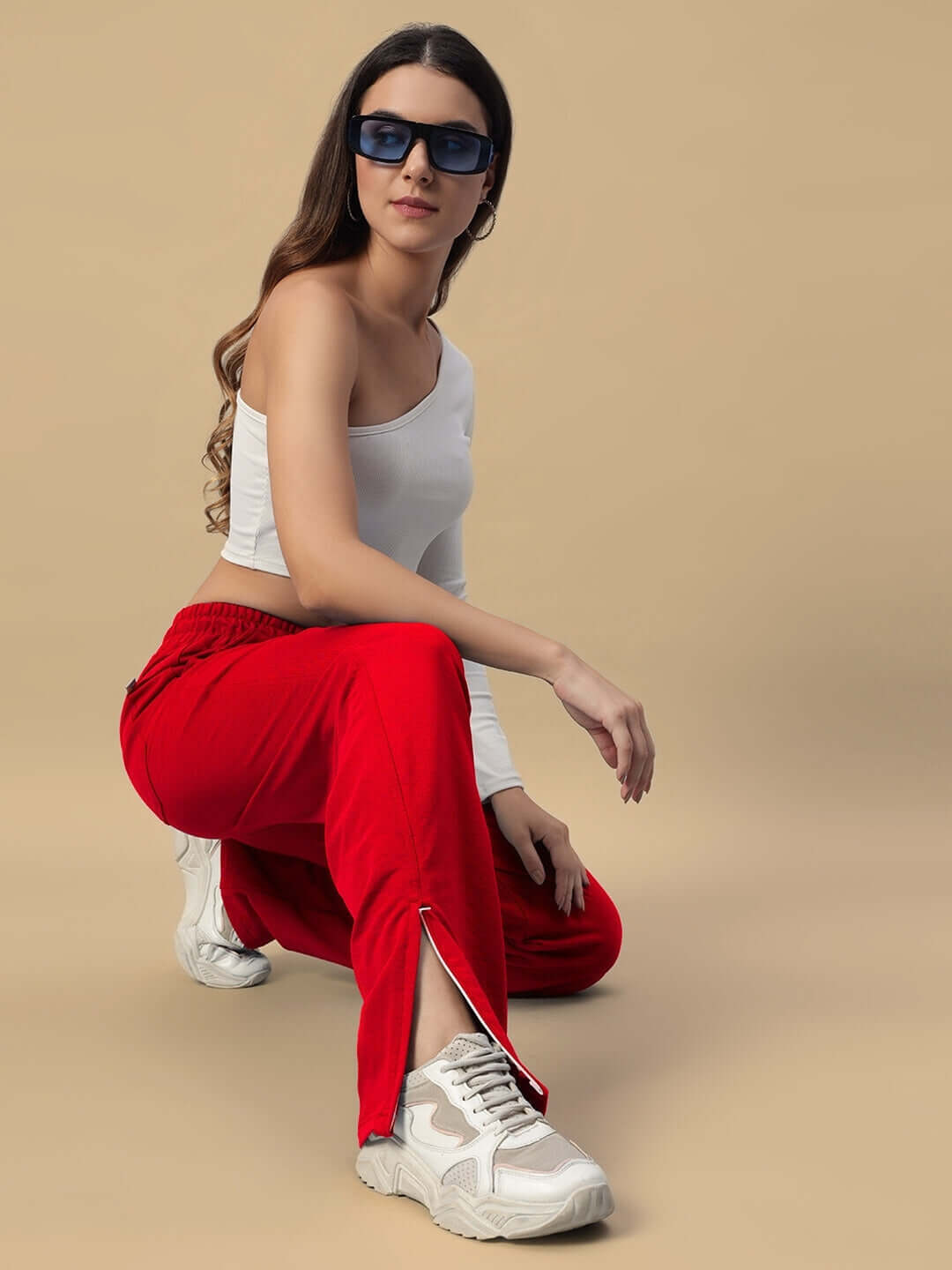 Woman wearing red Hershey side slit joggers and sunglasses, crouching in a stylish pose against a beige background.