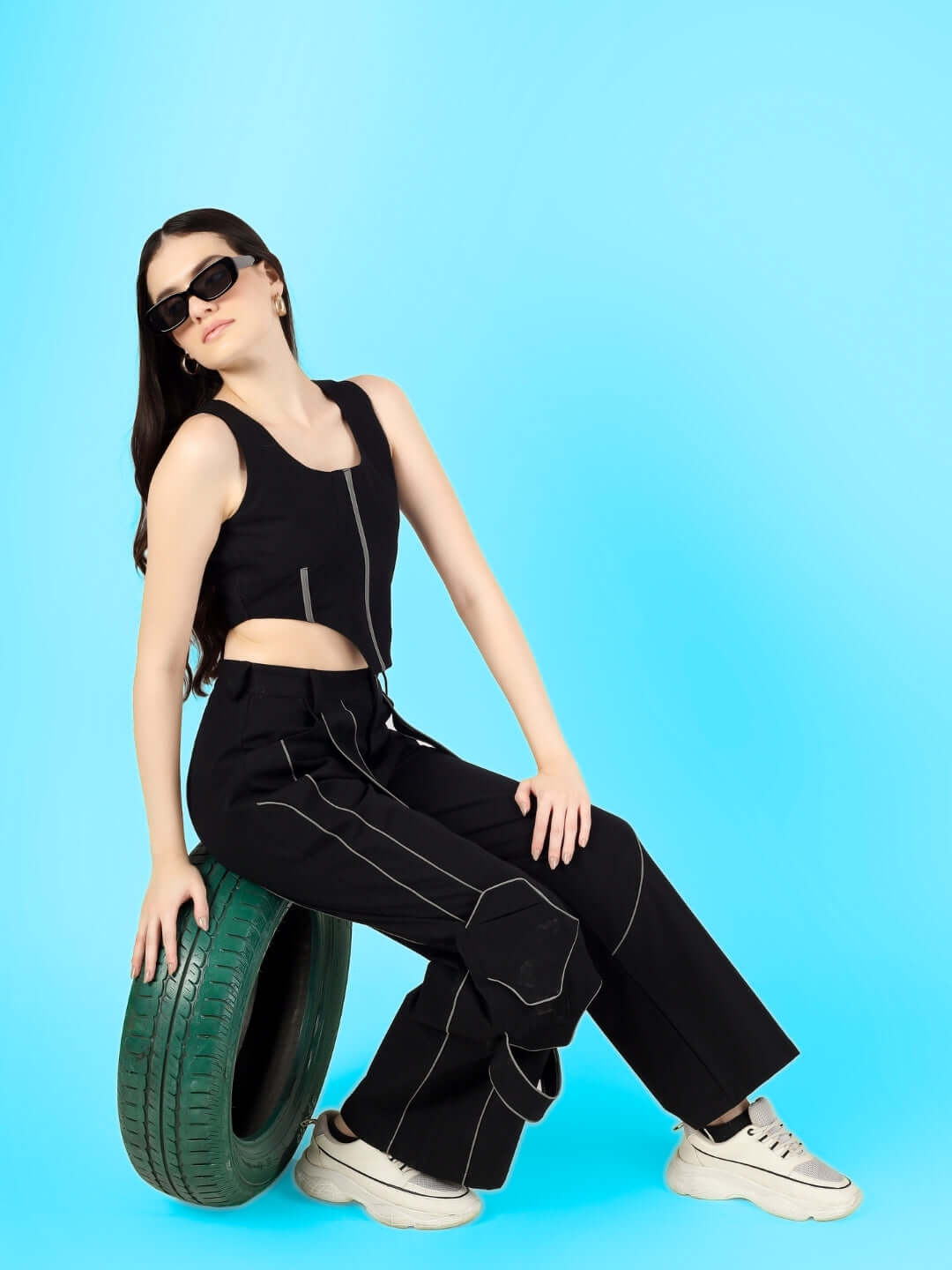 Woman wearing a stylish black reflective co-ord set with a corset top and carpenter pants, sitting on a tire against a blue background.