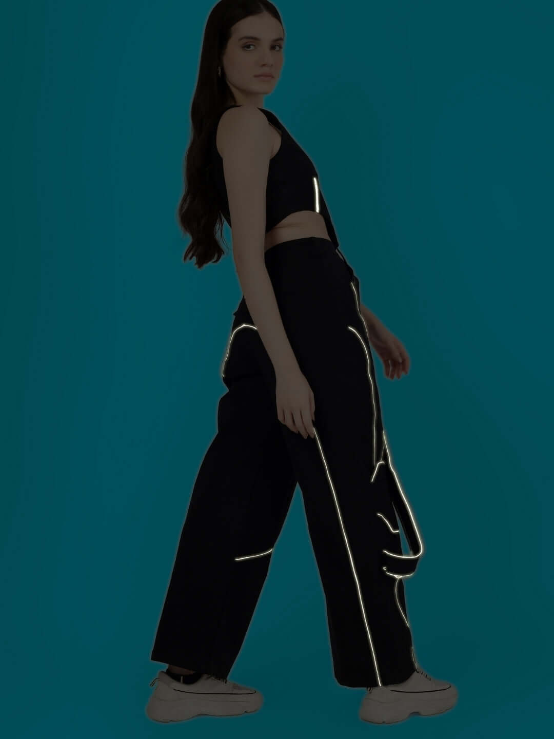 Model wearing women's reflective co-ord set with corset top and reflective carpenter pants by DUDS on a teal background.