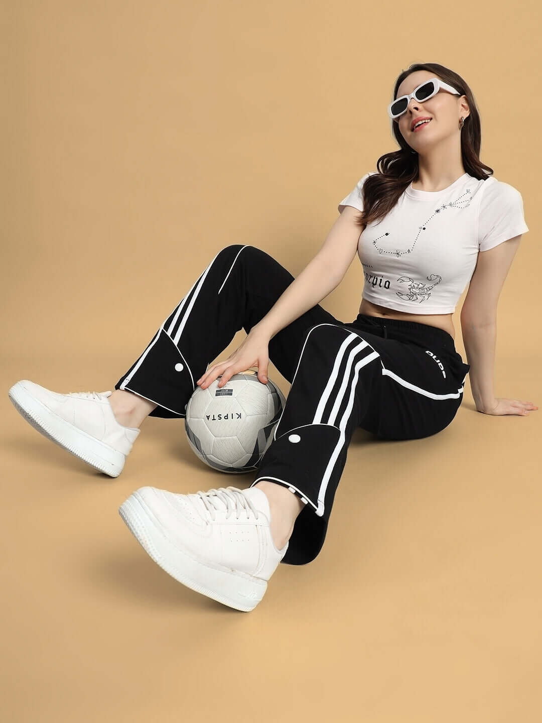 Woman wearing WOMEN SCORPIO CROP-TOP BABY PINK sitting on floor with soccer ball, dressed in sporty attire and sunglasses.