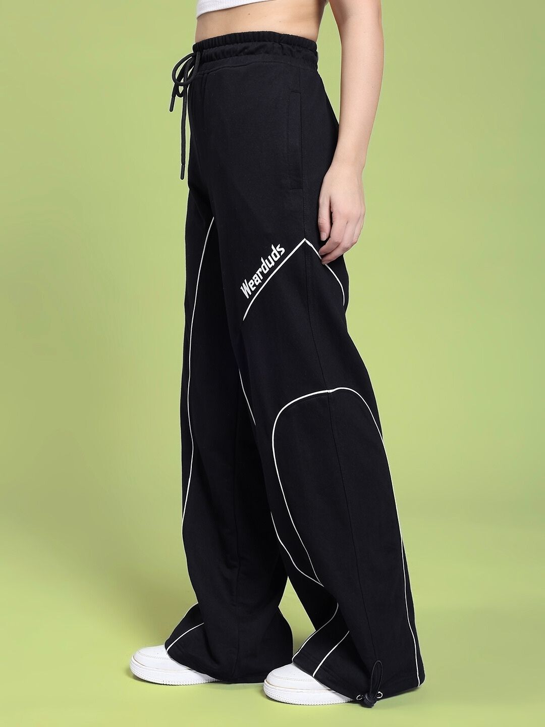 Side view of WOMEN SIGNATURE PIPING JOGGERS BLACK with contrast piping, showcasing street-ready style against a green background.