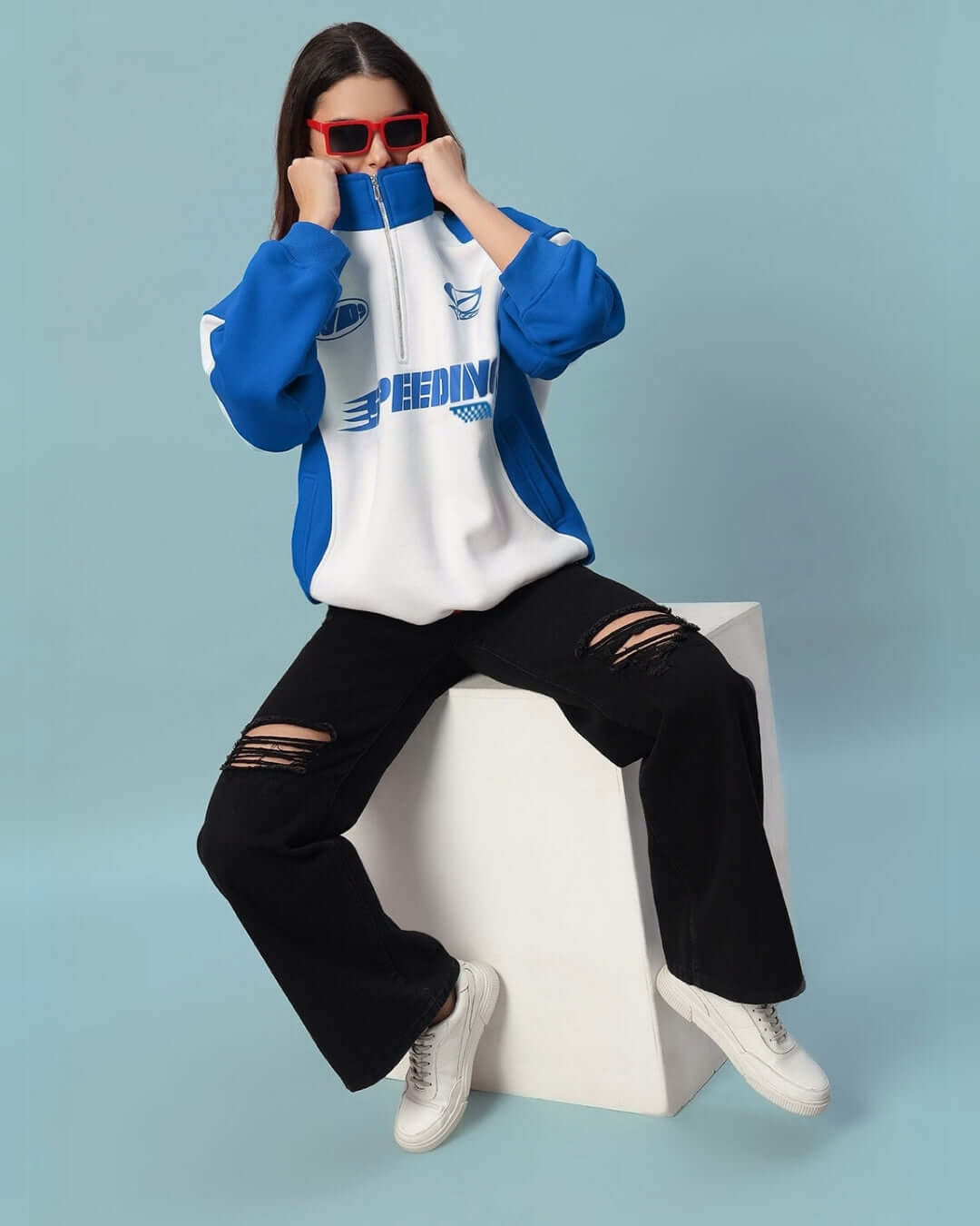 Woman wearing Women's Speeding Colorblock Jacket in royal blue with sporty design, ripped black jeans, and seated on a white block.