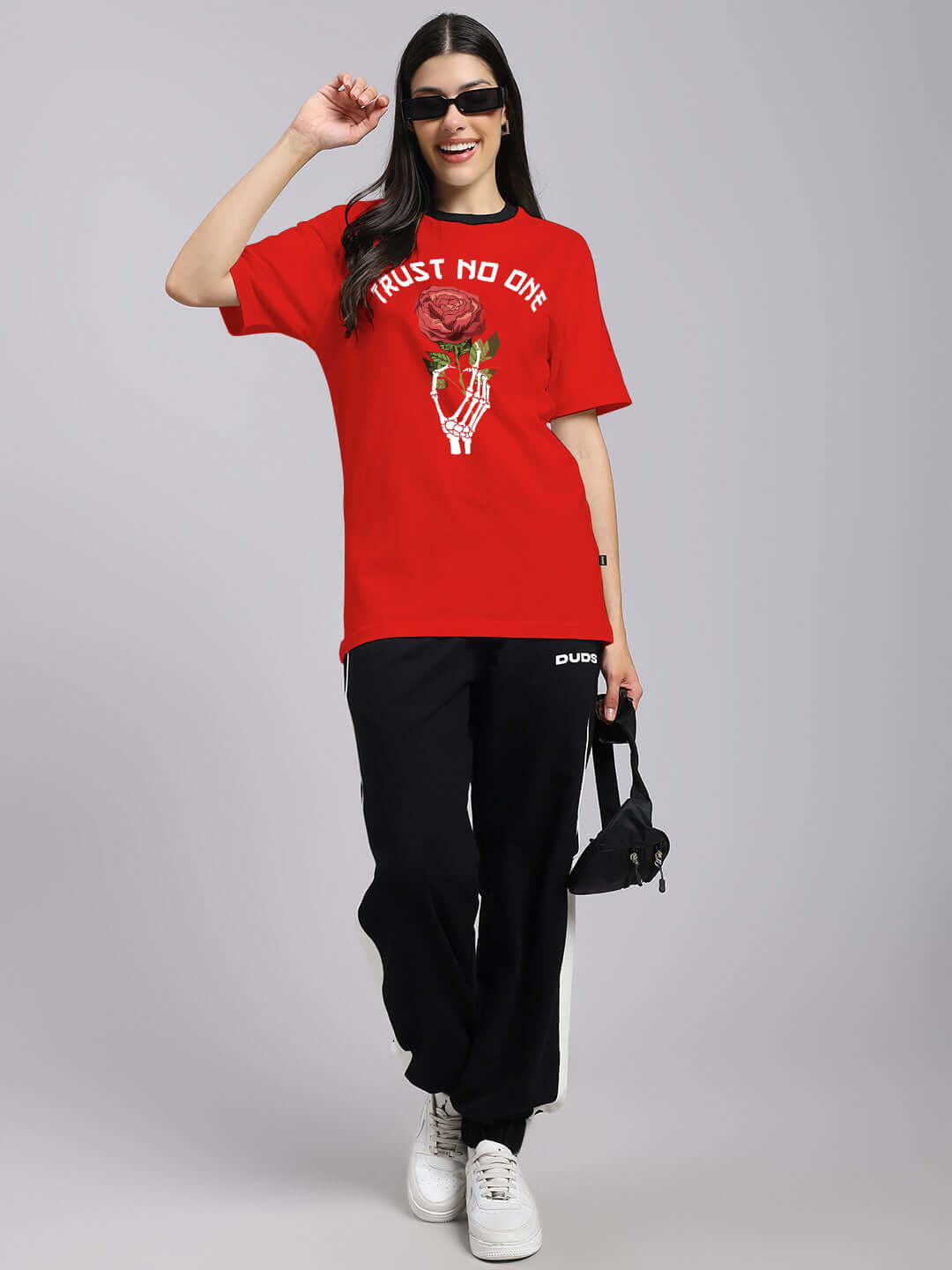 Woman in red 'Trust No One' printed t-shirt and black pants, wearing sunglasses, holding a black bag. Casual fashion statement.
