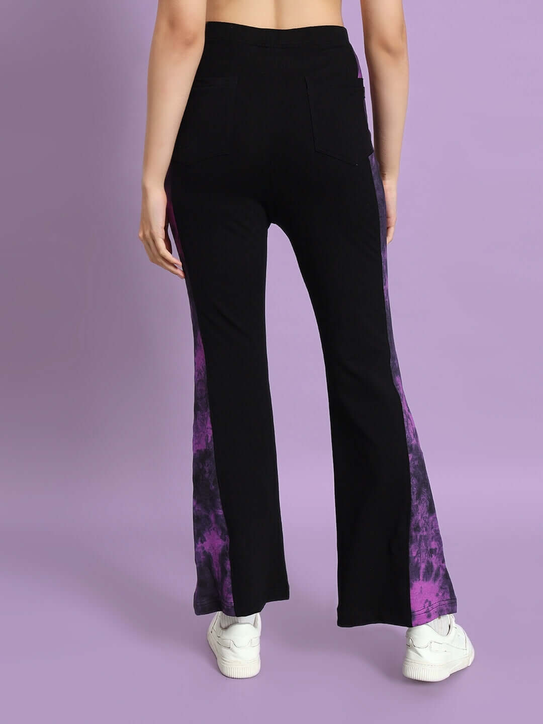 Back view of women's black and purple tie-dye panel boot cut pants worn by model, highlighting design and fit, against a purple background.