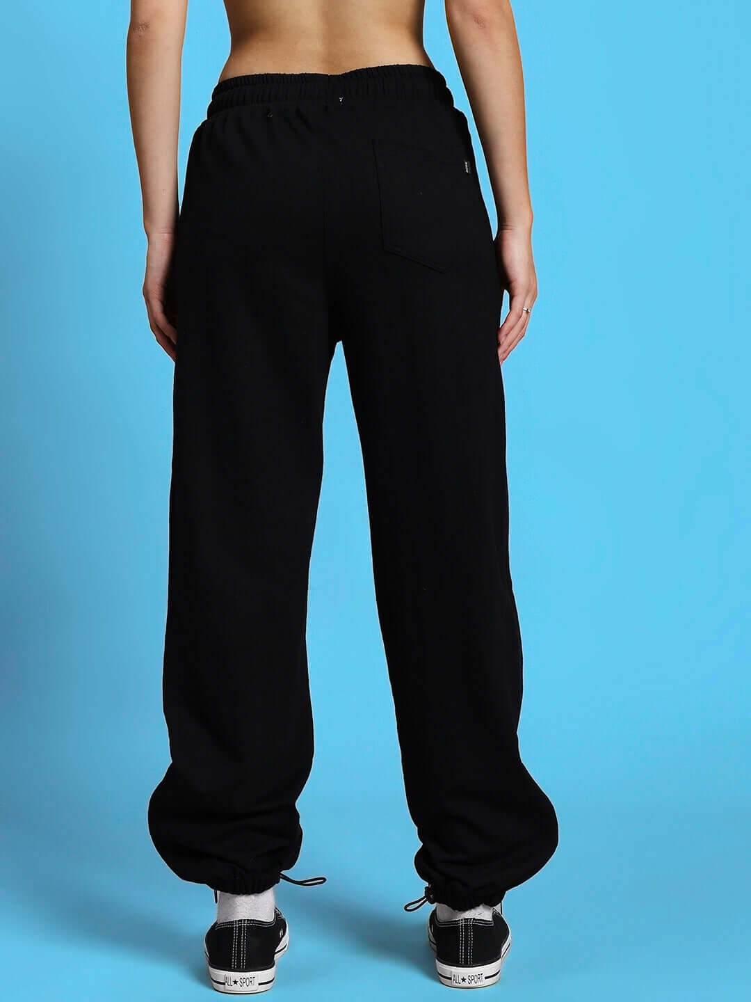 Back view of women's black zipper panel joggers with elastic waistband, worn with sneakers, against a blue background.