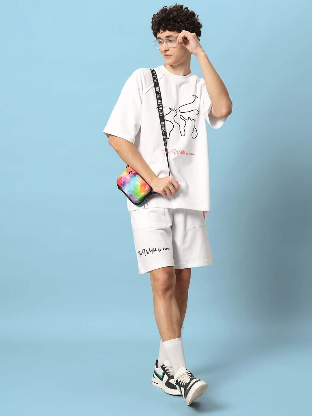 Model wearing 'The World is Mine' white printed co-ord set, featuring shorts and a t-shirt, accessorized with a colorful crossbody bag.