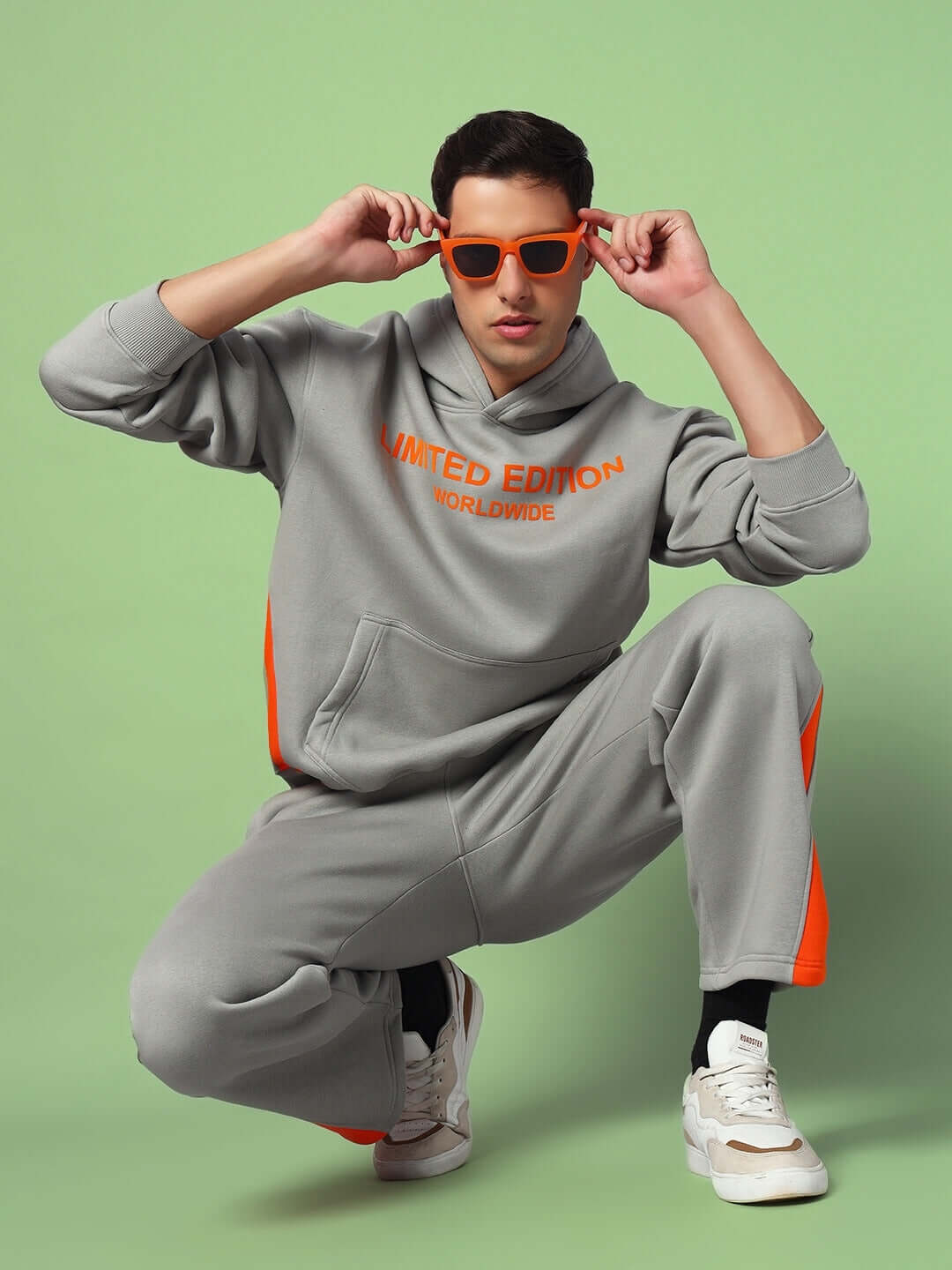 Man wearing WORLDWIDE FLEECE COLORBLOCK CO-ORD GREY with orange accents, posing in stylish sunglasses against a green background.