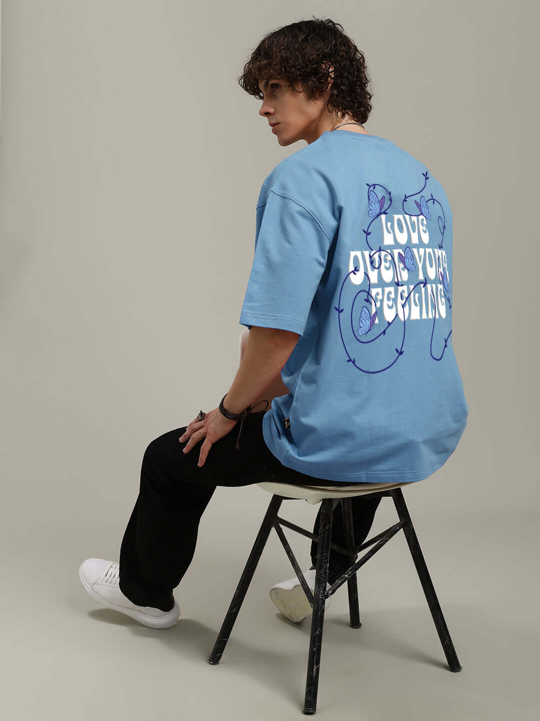 Model wearing a light blue printed T-shirt with 'Love Over Your Feelings' design, made from premium cotton blend, sitting on a stool.