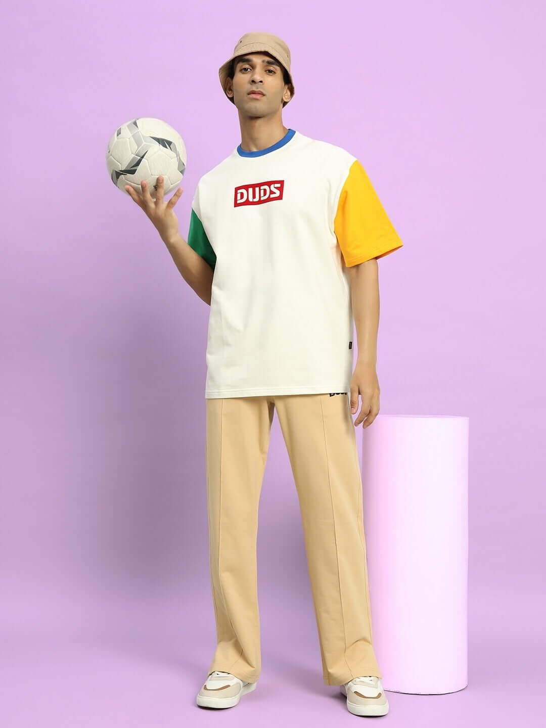 Model wearing beige Zion Side Slit Joggers by DUDS, paired with a colorful t-shirt, holding a soccer ball against a purple background.