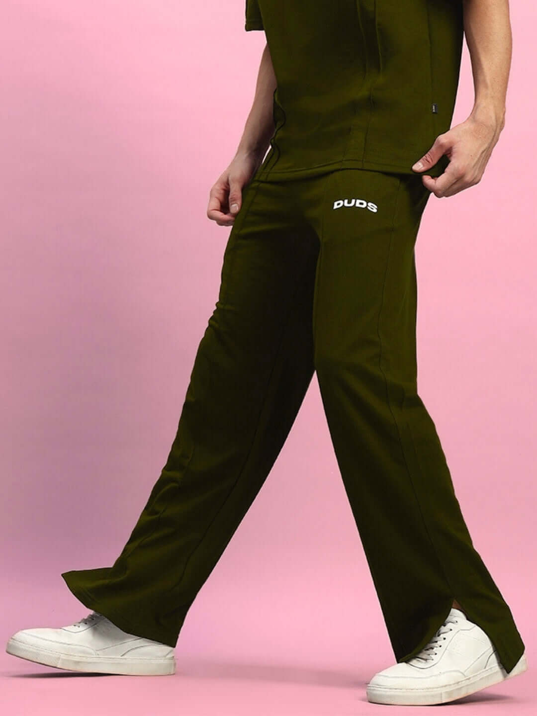 Olive green side slit joggers by DUDS, shown from the side, worn with white sneakers against a pink background.