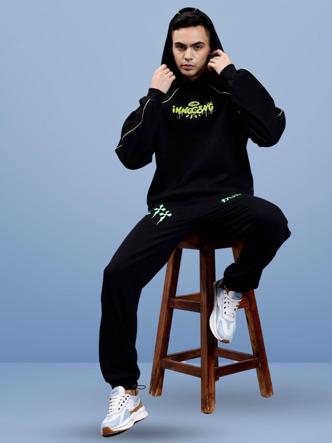 Model wearing black hoodie and joggers with neon accents, sitting on a wooden stool against a blue background.
