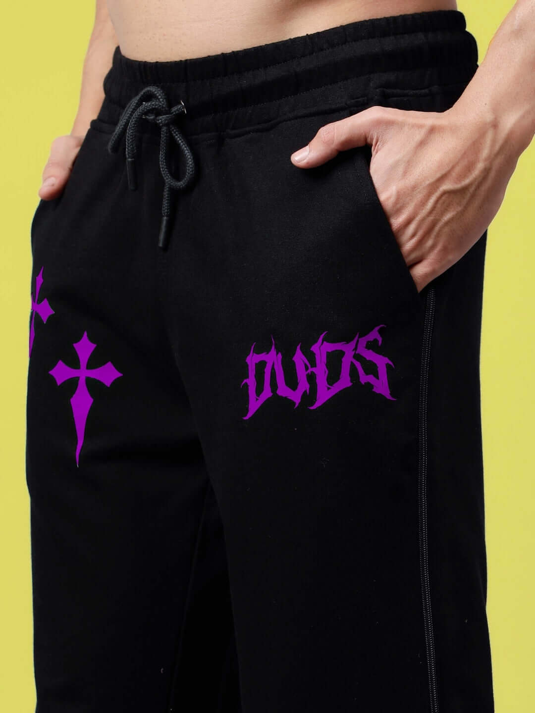 Close-up view of purple zipper panel joggers with bold graphics, showcased on a model's waist against a vibrant yellow background.