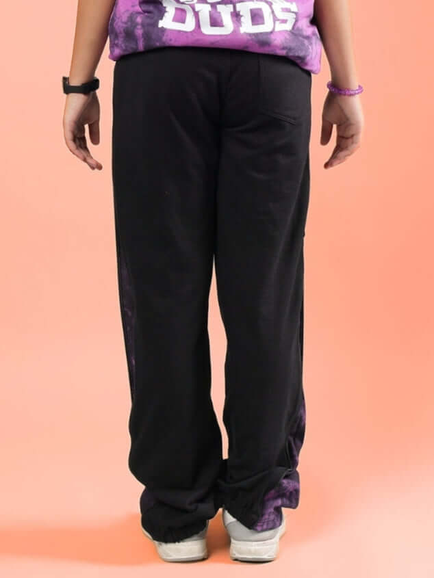 Child wearing black and purple zipper panel jogger pants from DUDS, viewed from the back, on a peach background.